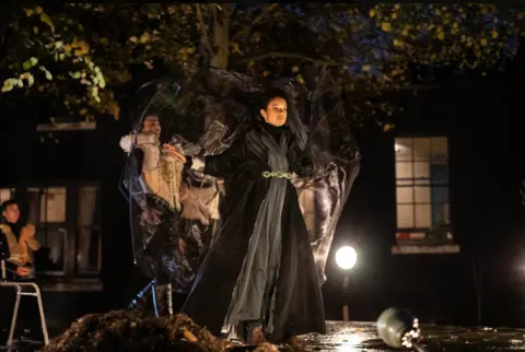 Joseph Winters Woman in dark cape on stage in front of spotlights