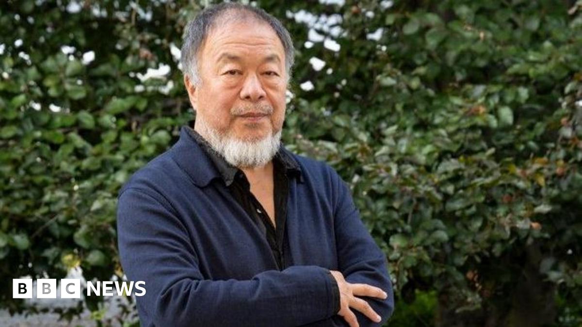 Ai Weiwei, who is wearing a black shirt and a navy overshirt, stands in front of a hedge with his arms crossed. He has a grey beard.