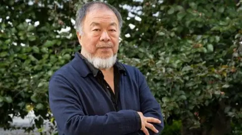 Getty Images Ai Weiwei, who is wearing a black shirt and a navy overshirt, stands in front of a hedge with his arms crossed. He has a grey beard. 