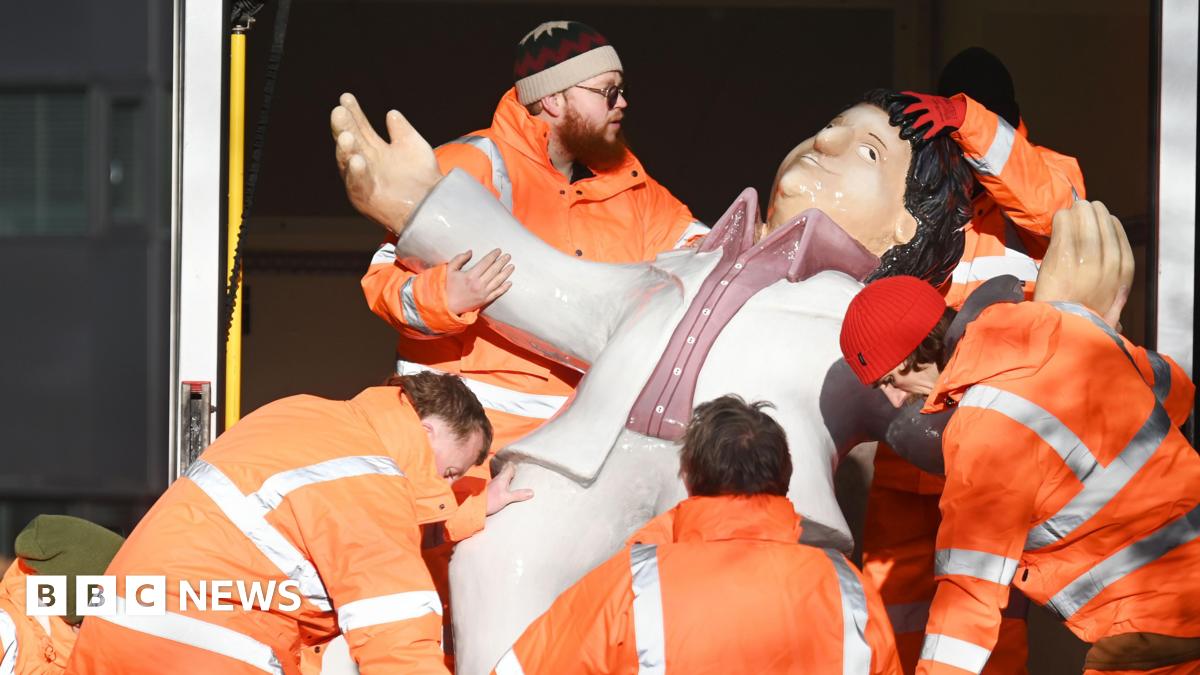 Five men in high viz orange jackets, carrying one of Beryl Cooks sculptures out of a van. The sculpture is of 'Dancing Tom'.