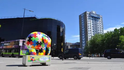 A multi-coloured Elmer sculpture on a pavement with the dark glass-fronted Willis Building and a residential tower block in the background on the other side of a road, which has a bus and two other vehicles on it.