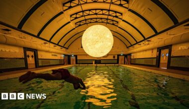 A silhouette of a swimmer diving into an indoor pool. There is a large sculpture modelled off of the sun suspended above the pool. It is reflecting in the water.