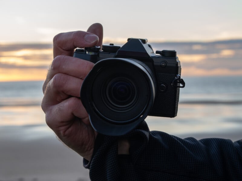 A close-up of a hand holding a black camera, with the lens pointing toward the viewer. In the background, there is a blurred beach scene at sunset, with soft light and clouds in the sky.