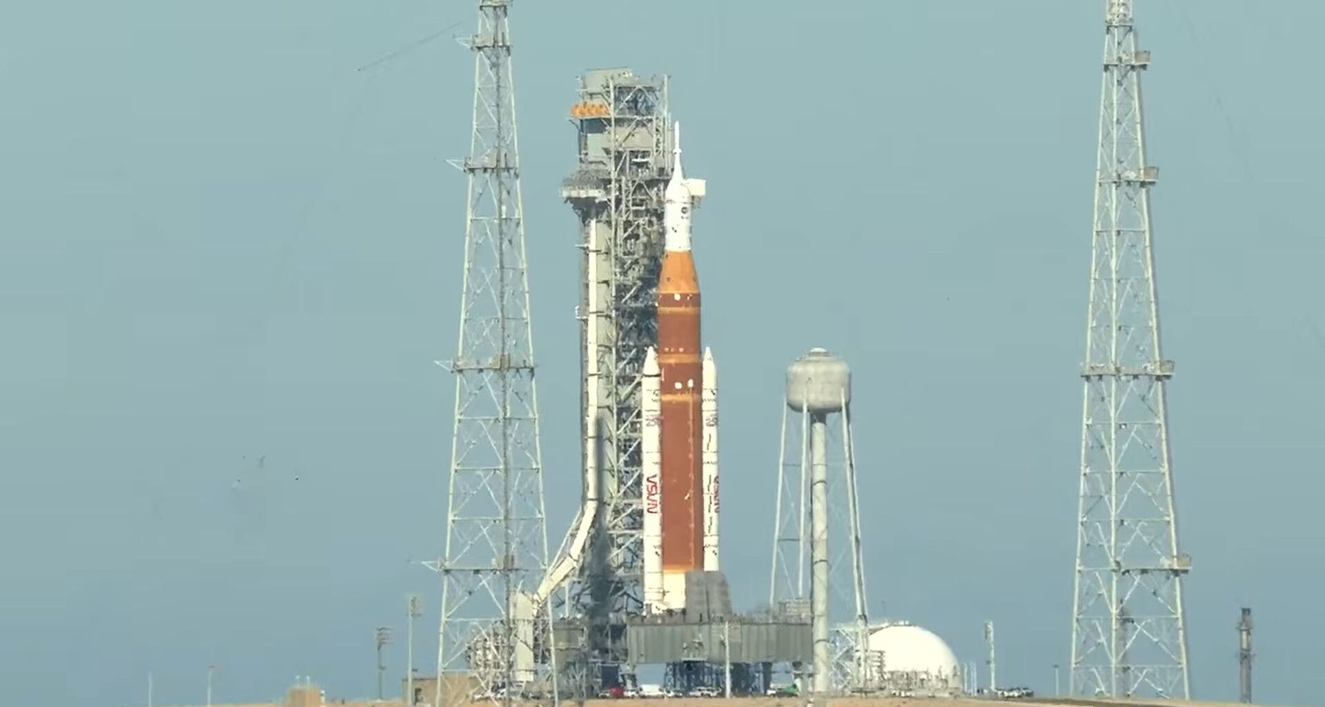An orange and white rocket is held vertically by scaffolding on a launch pad.