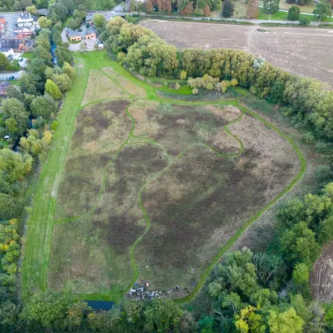 Matt Elson An aerial view shows a large, roughly heart‑shaped area of open grassland bordered by dense trees. Winding narrow paths cut through the grassy field, creating looping shapes across the middle. The grass varies in colour, with some patches looking brown with shrubs and others greener. To the left side of the image are houses and a small cluster of buildings surrounded by trees. On the right and upper edges of the image are open, ploughed fields. At the bottom edge, there is a small pile of pots and gardening equipment near the tree line. The landscape appears quiet and rural, with a patchwork of natural and managed spaces.