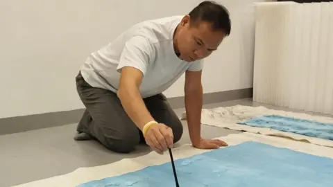 The Wilson Albert, who wears grey trousers, a white t-shirt and holds a long paintbrush, kneels over a large piece of paper which has been painted light sky blue. 