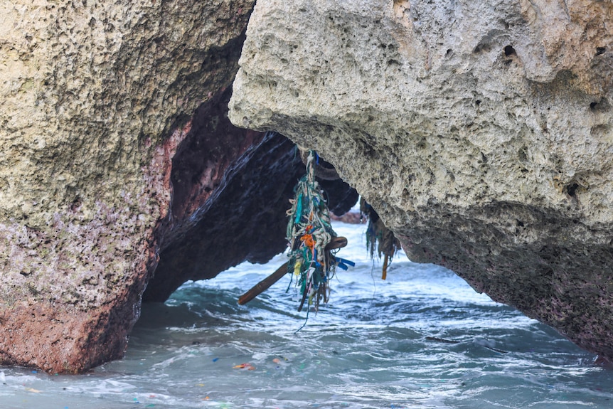 A rope with plastic and rubbish hanging off it lies above the water in between two large rocks.