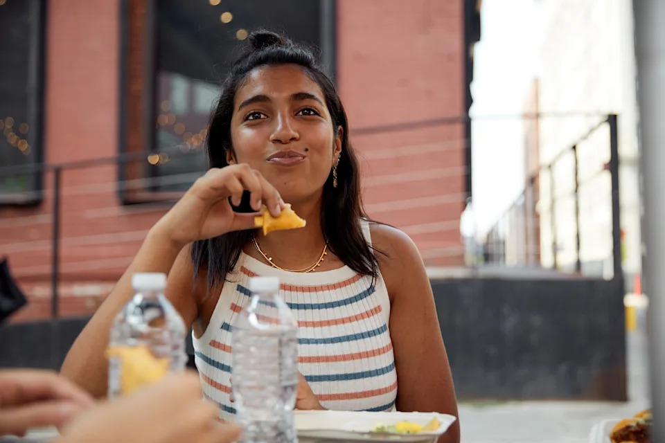 Person smiling while eating food outdoors, holding a piece of bread. There are water bottles and food containers on the table