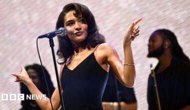 Olivia Dean wears a black dress and is performing on stage in front of a microphone. She is looking off to the side and pointing outwards with her hands.