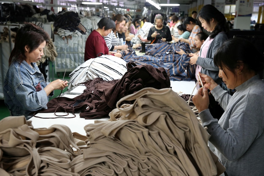 A row of Chinese women stand at a long table, sewing from piles of fabric and clothing.