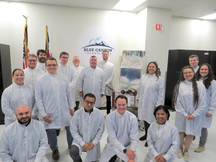 A group of people wearing lab coats, standing next to a satellite.