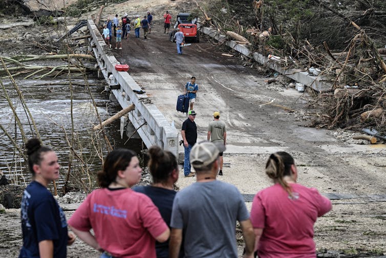 People stand near a bridge and searchers look through debris that has washed up.