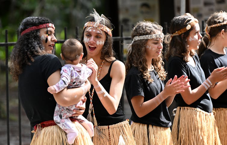 A line of women, including one holding a baby, participate in a cultural event dressed in First Nations clothing.