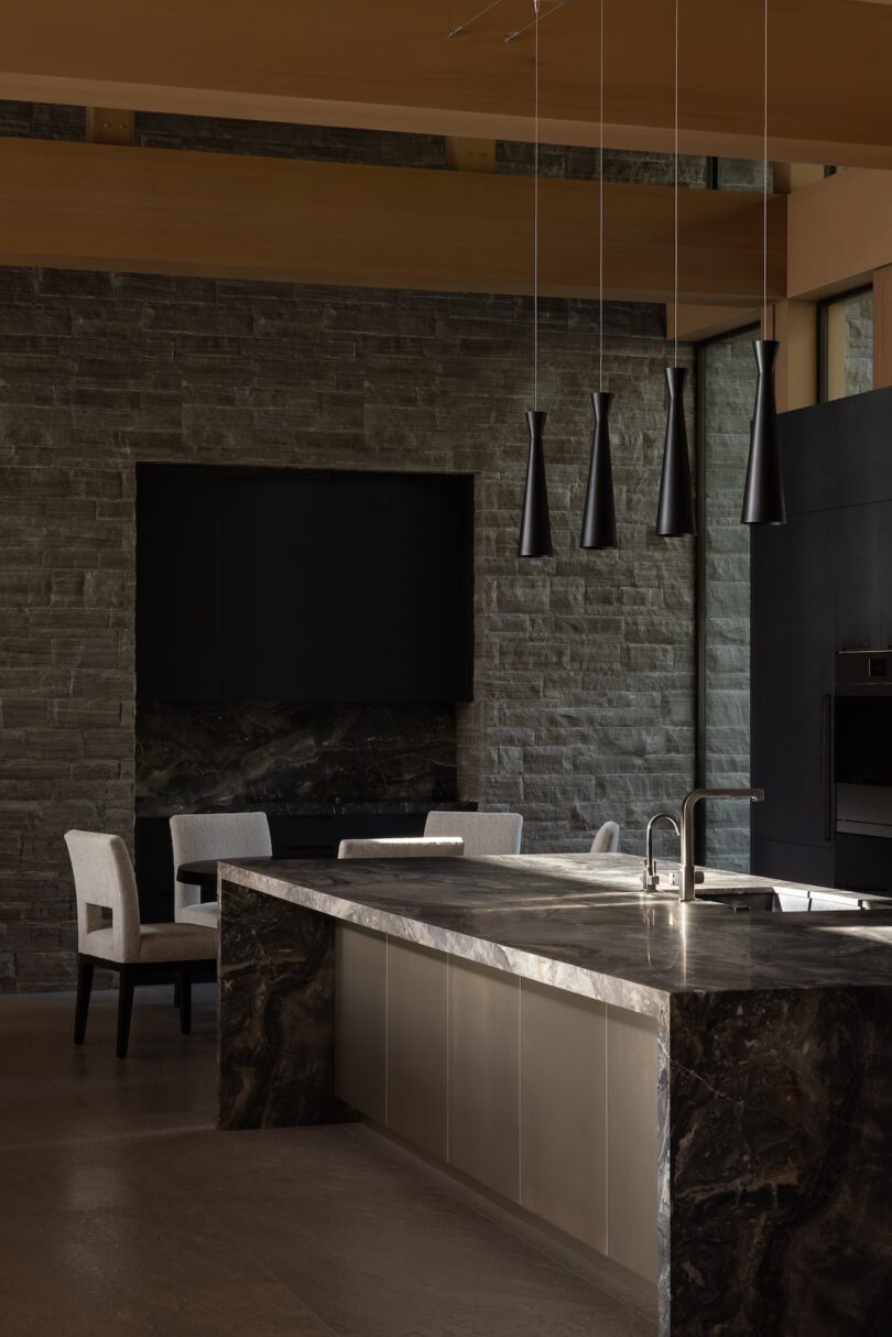 Modern kitchen with a marble island, built-in sink, four pendant lights, stone walls, and two upholstered chairs by a dark fireplace.