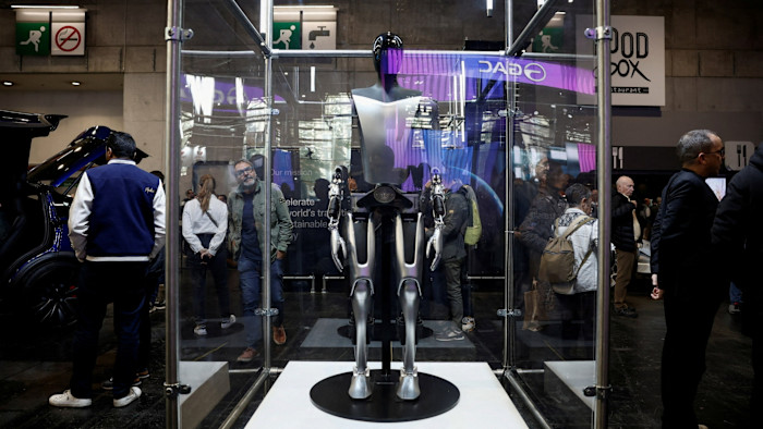 The Tesla Optimus robot is displayed inside a glass case at the 2024 Paris Auto Show, with people observing around it.