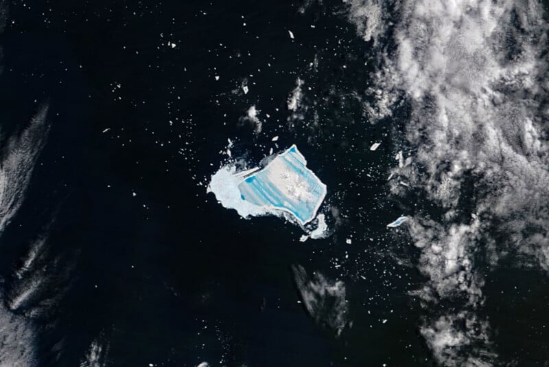 Aerial view of a large iceberg with bright blue stripes floating in dark ocean water, surrounded by smaller ice fragments and patches of white clouds.