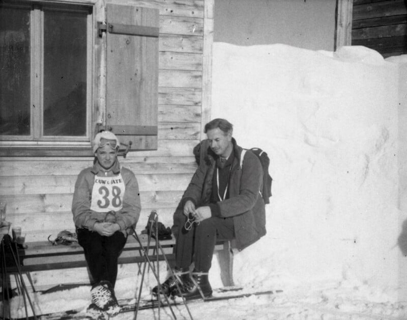 Two people sit on a wooden bench outside a snow-covered cabin. One wears ski gear and a bib with the number 38, while the other adjusts ski equipment. Snow is piled high beside the wooden building.