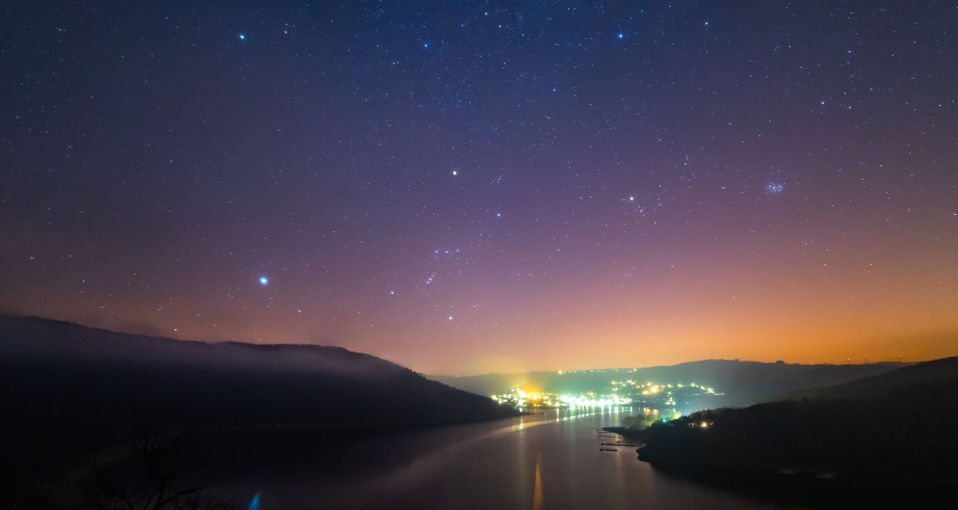 A purple and orange night sky with a few twinkling white stars overlooks distant city lights with a river and two dark banks in the foreground