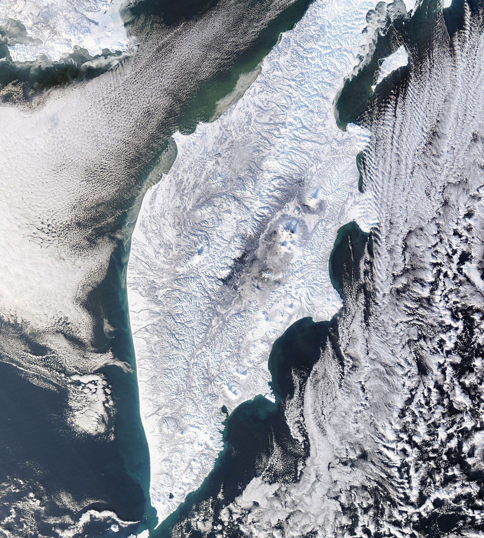 A thick layer of white snow blankets the Kamchatka Peninsula. Layers of clouds surround the peninsula, framing it but leaving its coastlines and a narrow portion of ocean visible around it. On land, several large, circular volcanoes dot the rugged landscape.