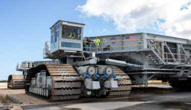 A crawler-transporter, including its massive treads take up the majority of the image. In fact, the entire crawler cannot be seen. There are a few people at the top of the crawler, which allows you to see just how massive the machine is.