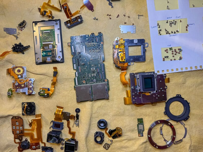 Camera parts are neatly laid out on a yellow cloth, including circuit boards, lenses, flex cables, screws, and a sensor. A piece of paper with sorted screws and handwritten labels is in the top right corner.