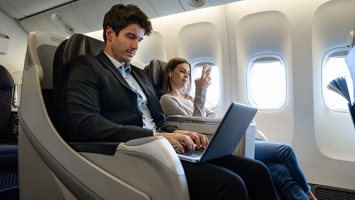 Airline passengers seated in an airplane cabin, with a man working on a laptop and a woman using a smartphone during a flight.