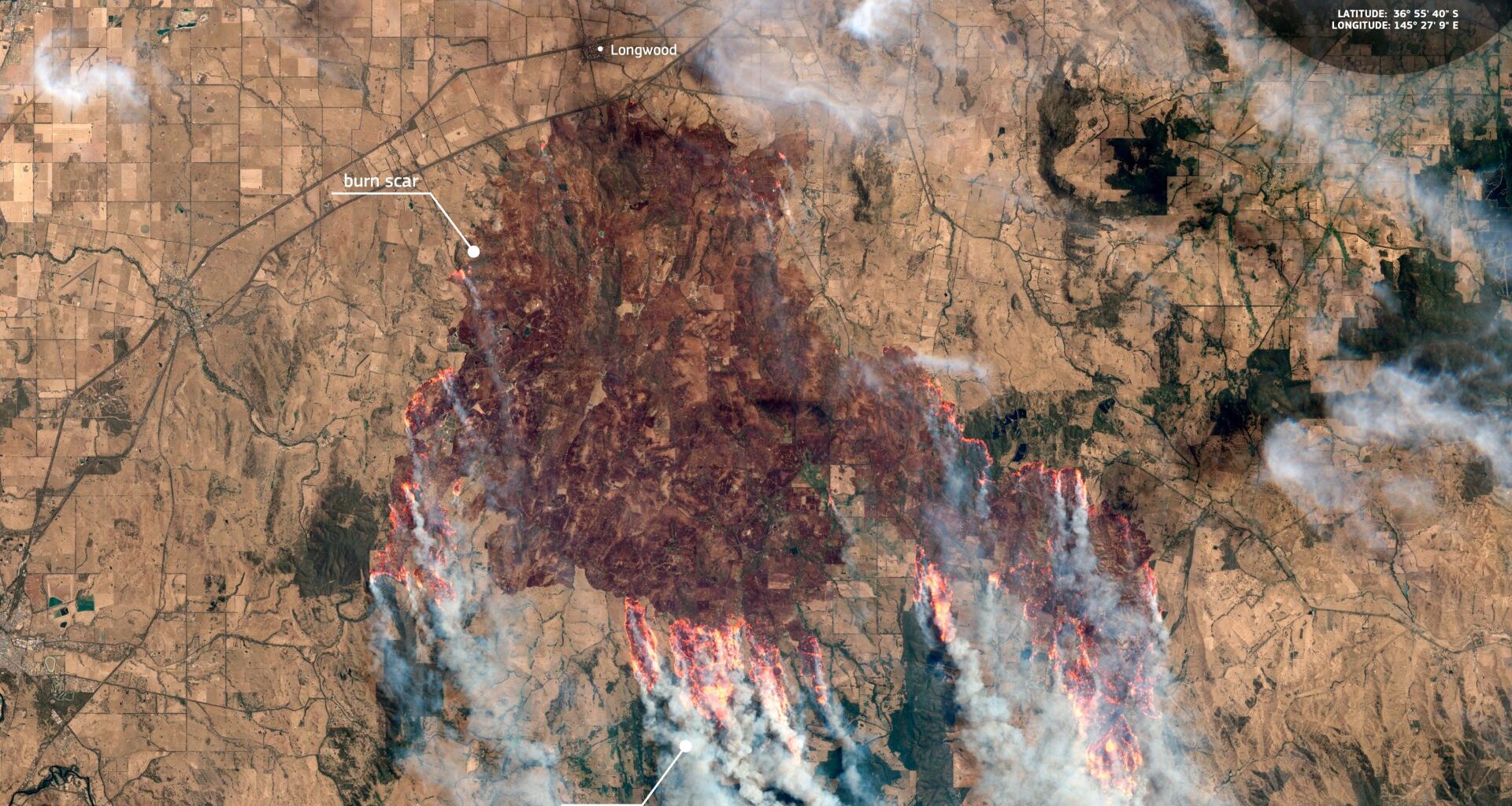 A brown landscape is scarred dark red as streaks of orange flames are seen around the edges of the dark area with streams of smoke moving toward the bottom of the image