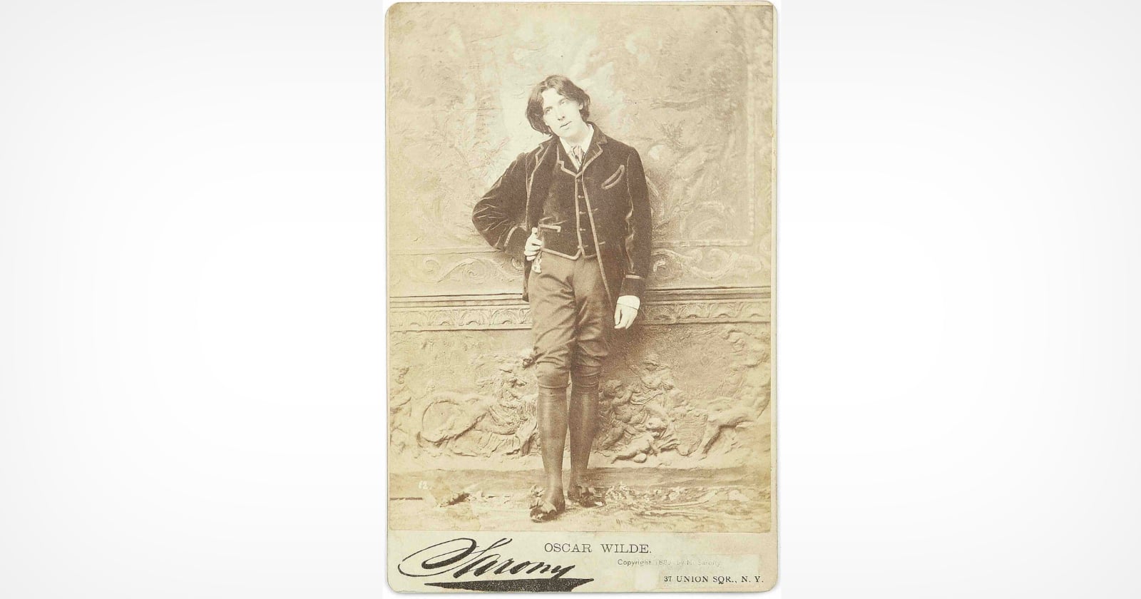 A sepia-toned portrait of Oscar Wilde standing and leaning against an ornate wall, dressed in Victorian-era clothing, with one hand on his hip and the other resting on the wall.