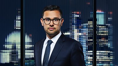 A man in glasses and a suit stands in front of a city backdrop 
