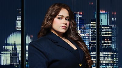 A woman with a nose piercing and long brown hair poses outside a city landscape at night 
