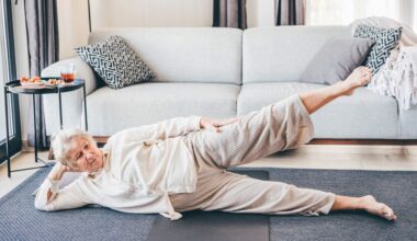 woman dressed in cream loose top and trousers lying on her side facing the camera with her top leg raised. she's on a grey carpet with a light grey sofa behind her.