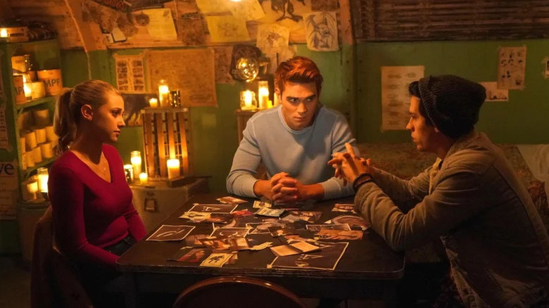 Lili Reinhart as Betty, KJ Apa as Archie and Cole Sprouse as Jughead sit at a photo-covered table in Riverdale
