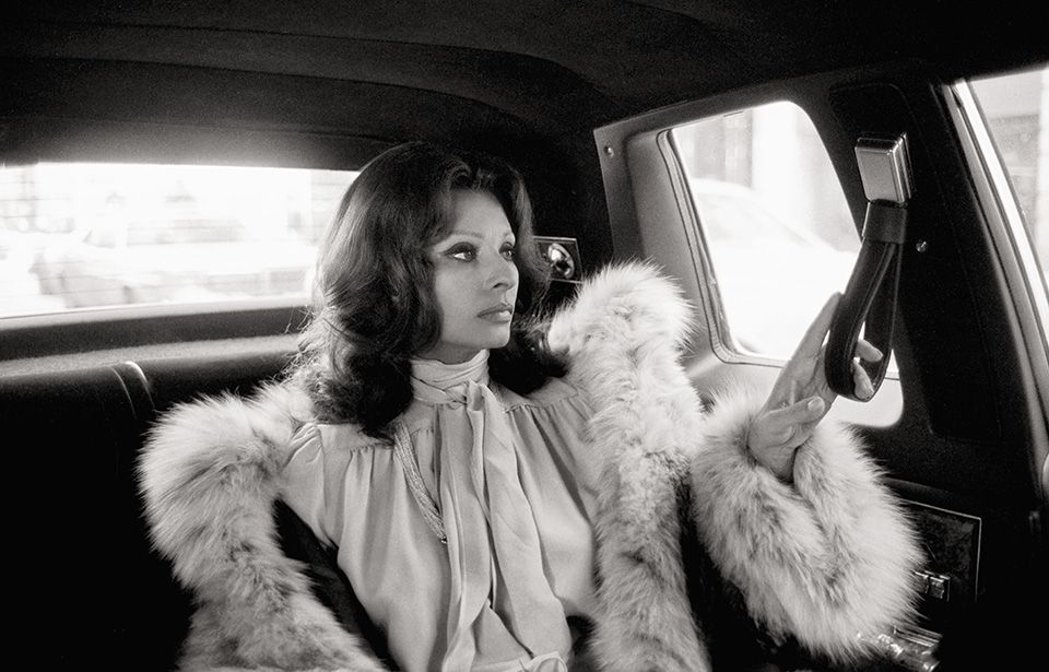 Sophia Loren is seen in a profile view sitting in the back of a car, wearing a voluminous fur-trimmed coat and a pussy-bow blouse.
