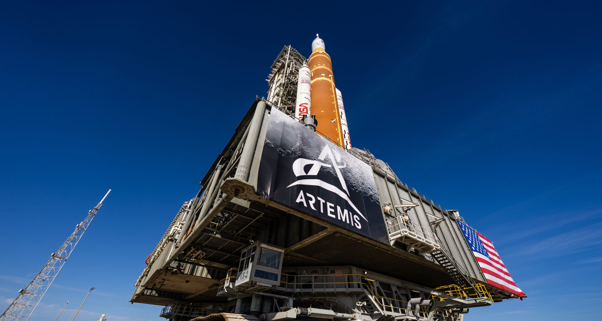 NASA's Artemis II SLS rocket - with its distinctive butterscotch orange-brown color- and Orion spacecraft are on top of a massive platform. The platform has a banner with the Artemis logo on it on the left and an American flag on the right. The rocket and platform are moved by an immense machine called the crawler-transporter, which has treads like a tank. The view is from the ground up, so the deep blue sky is the background for most of this picture.