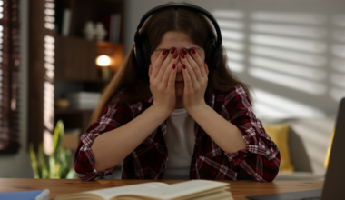 A young person with long hair covers their face in a distressed way while sitting at a desk with a book open in front of them.