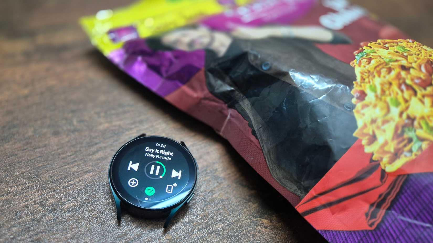 The Samsung Galaxy Watch 4 playing music on Spotify with a bag of snacks on the side.