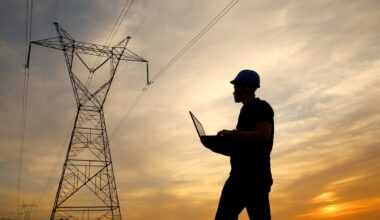 A person wearing a hardhat and holding a laptop near a power line.