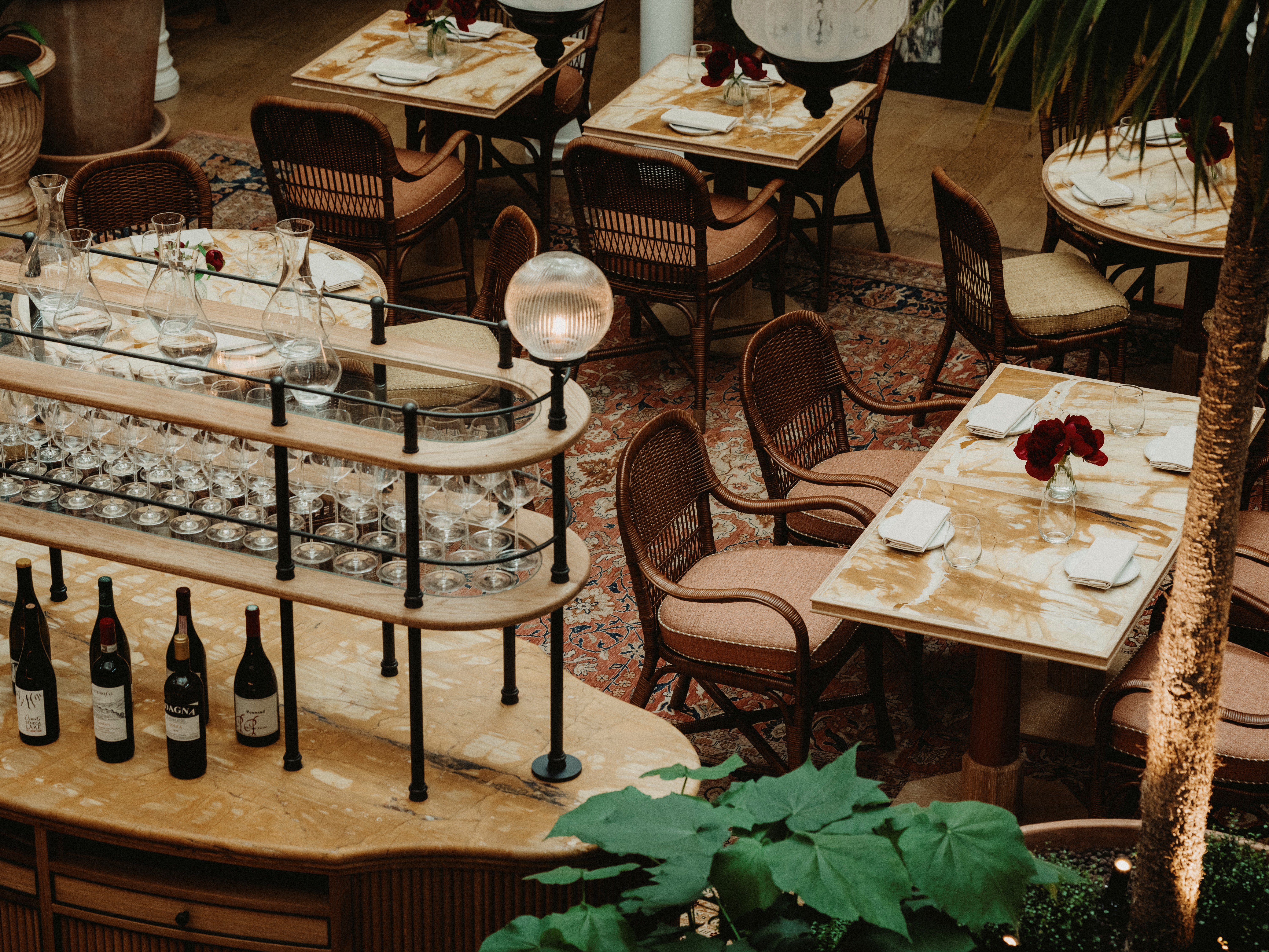 An Art Deco restaurant with marble-topped tables, wood and fabric chairs, patterned, red-toned rugs, and a central, oblong central bar with rounded lights.
