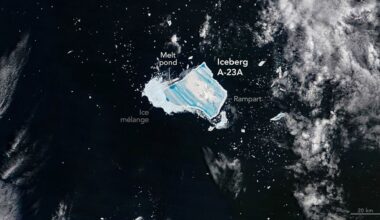 A blue jagged shape is seen in dark waters with white foam from the sea with the large white label Iceberg A-23A next to the blue shape