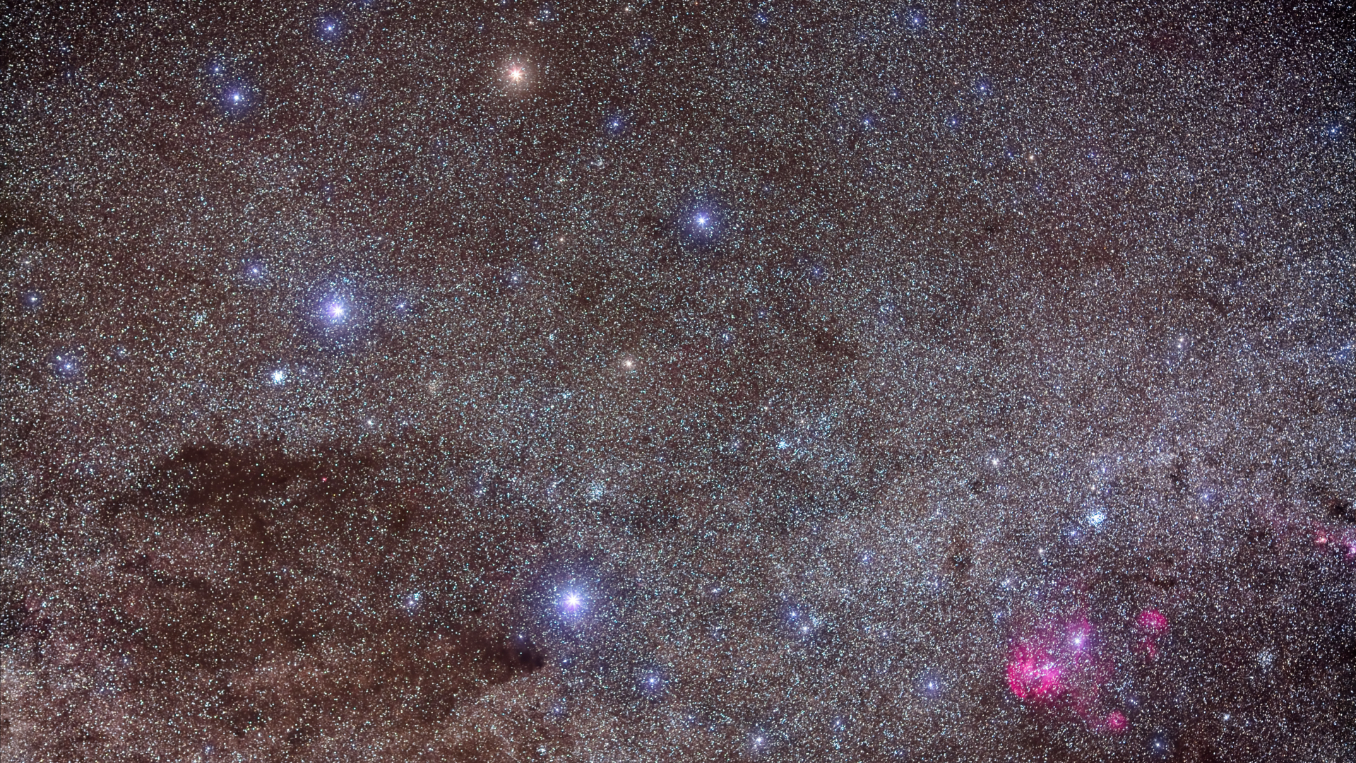 A glowing night sky full of stars with some bright blue and pink stars throughout with a dark area in the bottom left of the image