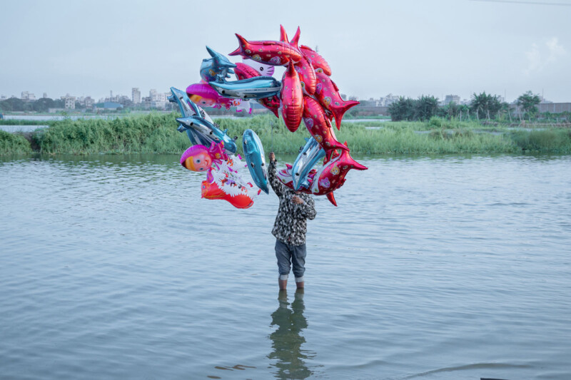 A person standing knee-deep in water holds a large bunch of colorful inflatable balloons shaped like fish and cartoon characters, obscuring their face and upper body. Green vegetation and distant buildings are visible in the background.