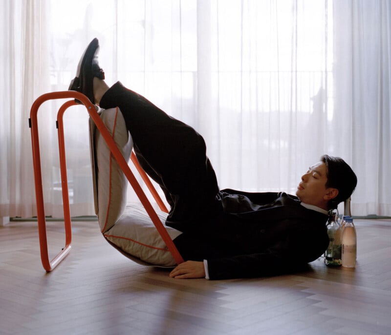 A man in a suit reclines on the floor with his legs raised on an orange metal frame. He appears relaxed, with his eyes closed, and there are bottles of water and a green bottle beside him on the wooden floor.