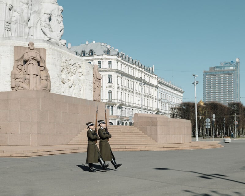 Two uniformed guards march in front of a large stone monument with sculptures, set in a mostly empty city square on a clear, sunny day. Historic and modern buildings line the background.
