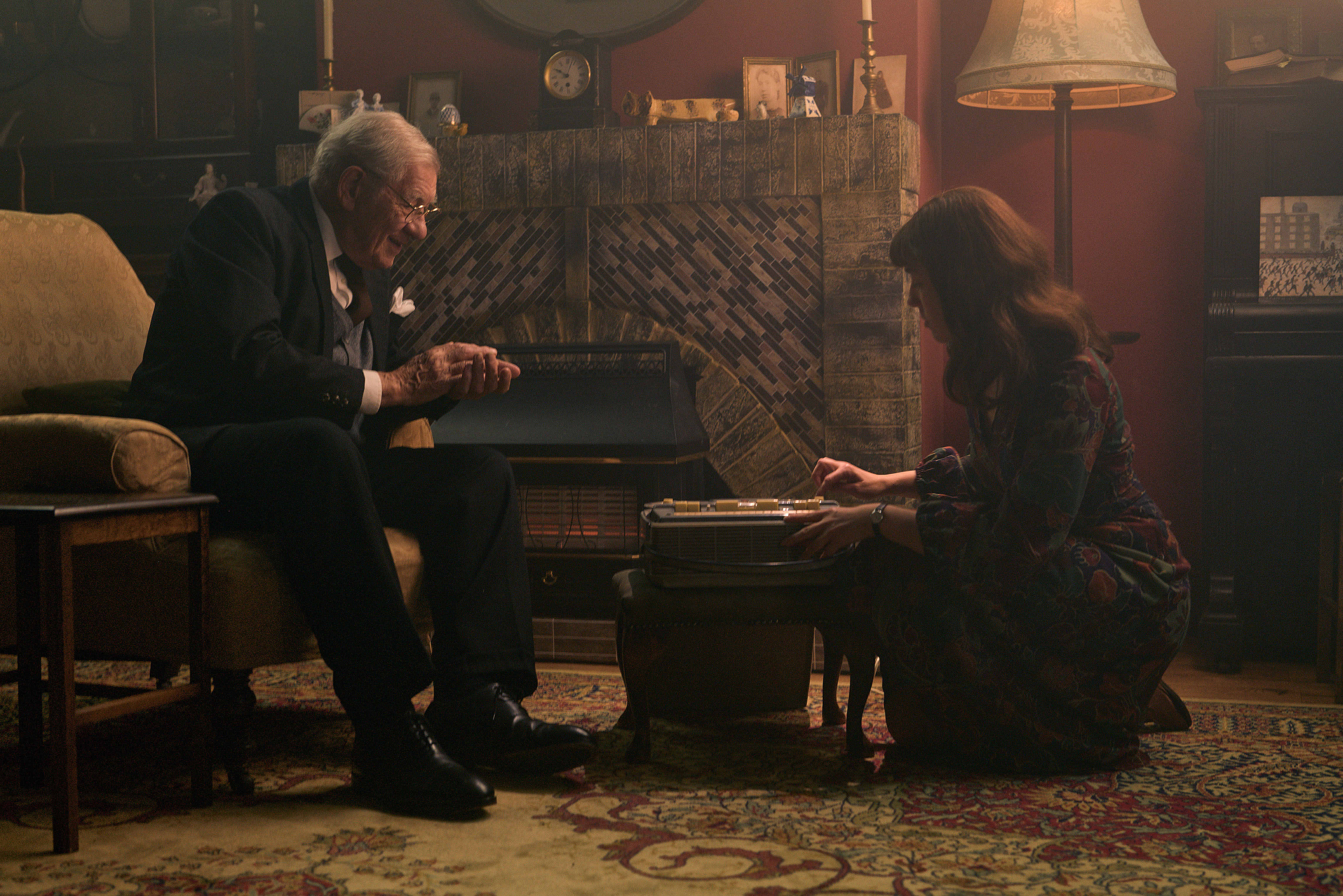 Ian McKellen as L.S. Lowry in a suit, sitting in a chair, conversing with Annabel Smith as Angela Barratt, who is kneeling by a tape recorder.