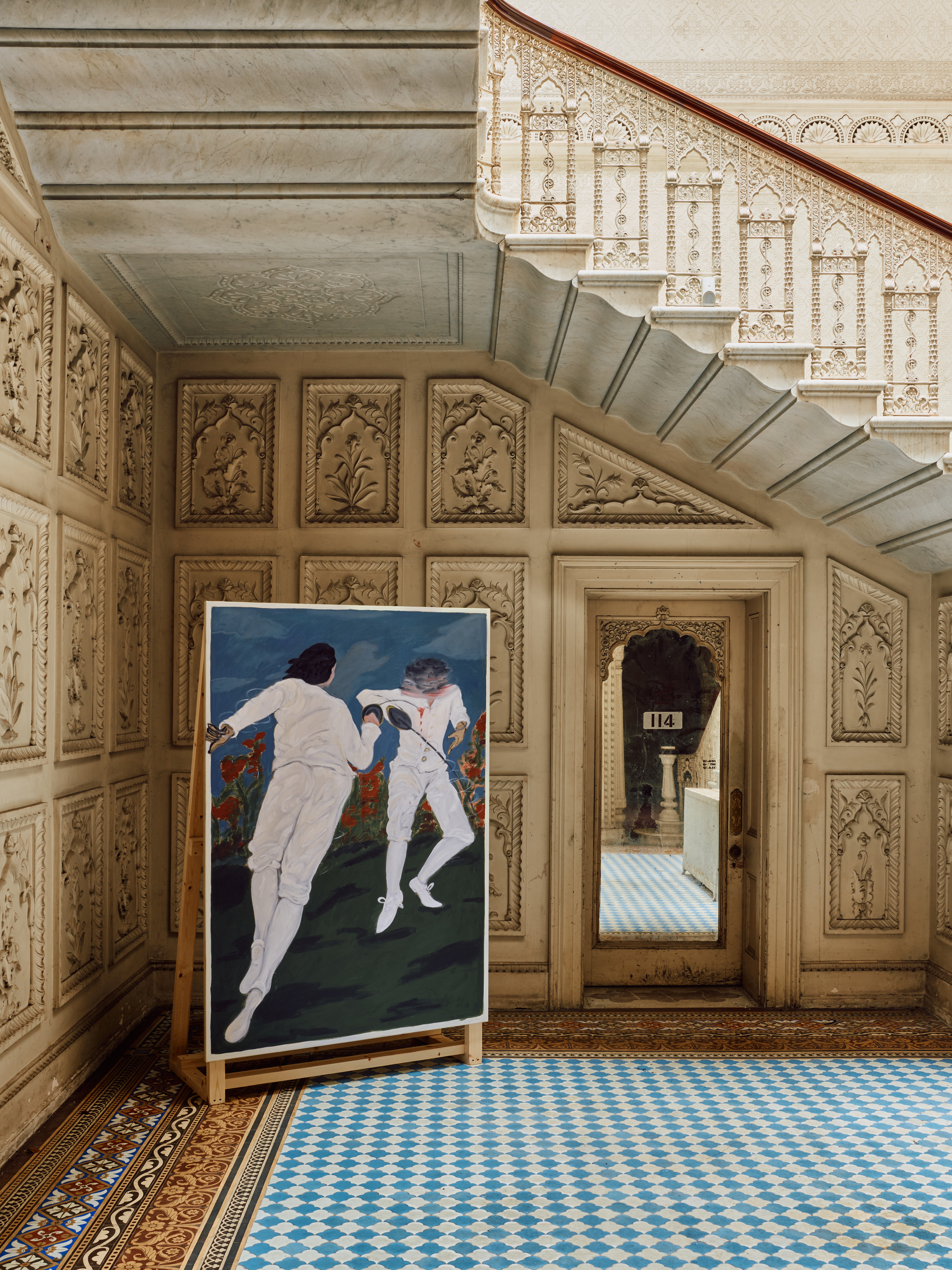 A large painting of two fencers in white stands on an easel in a room with elaborate white and gold carved walls, a white marble staircase with an ornate railing, and a patterned blue and white tile floor.