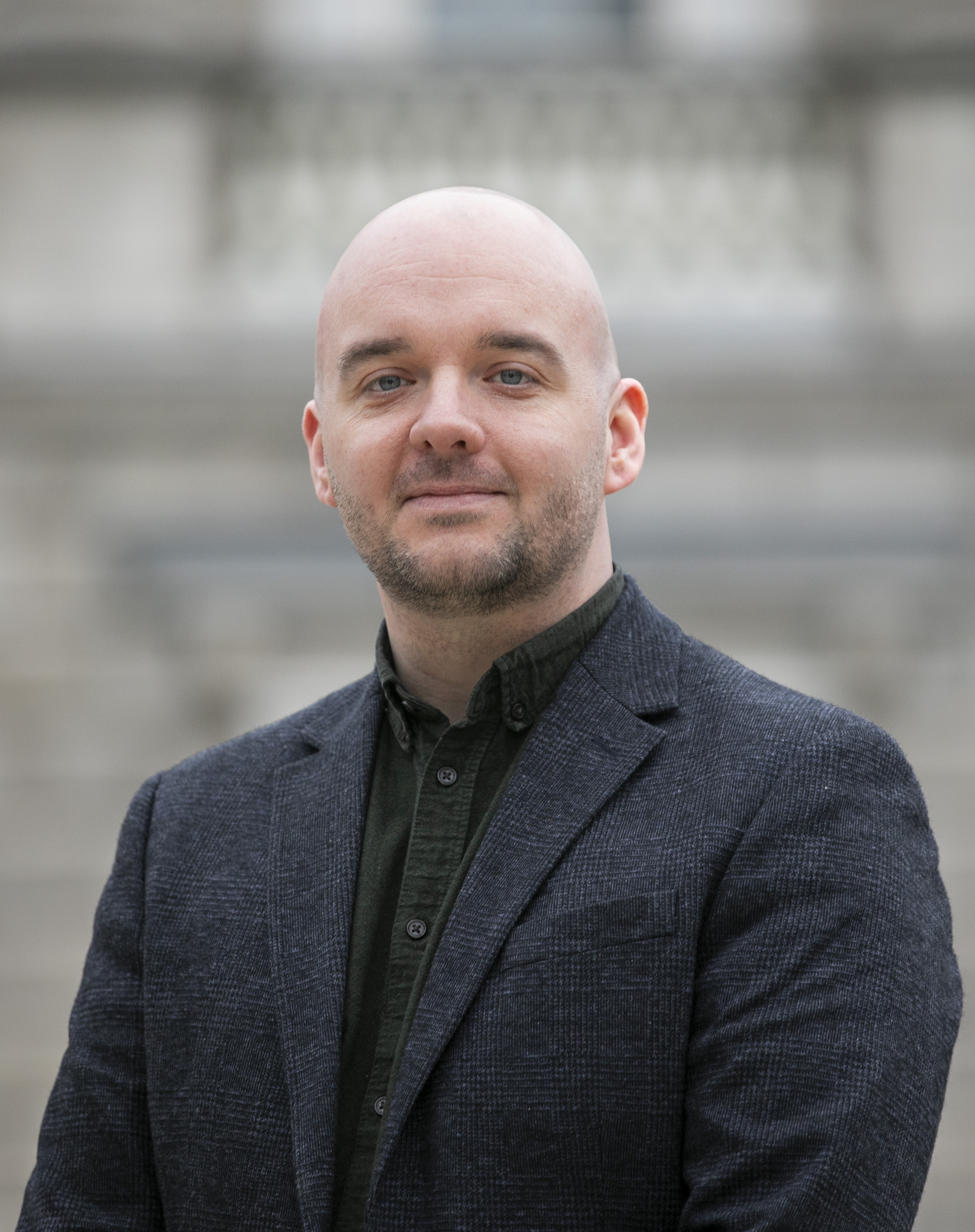 Aidan Farrelly, a man with a bald head and a beard, wearing a dark green shirt and gray blazer.