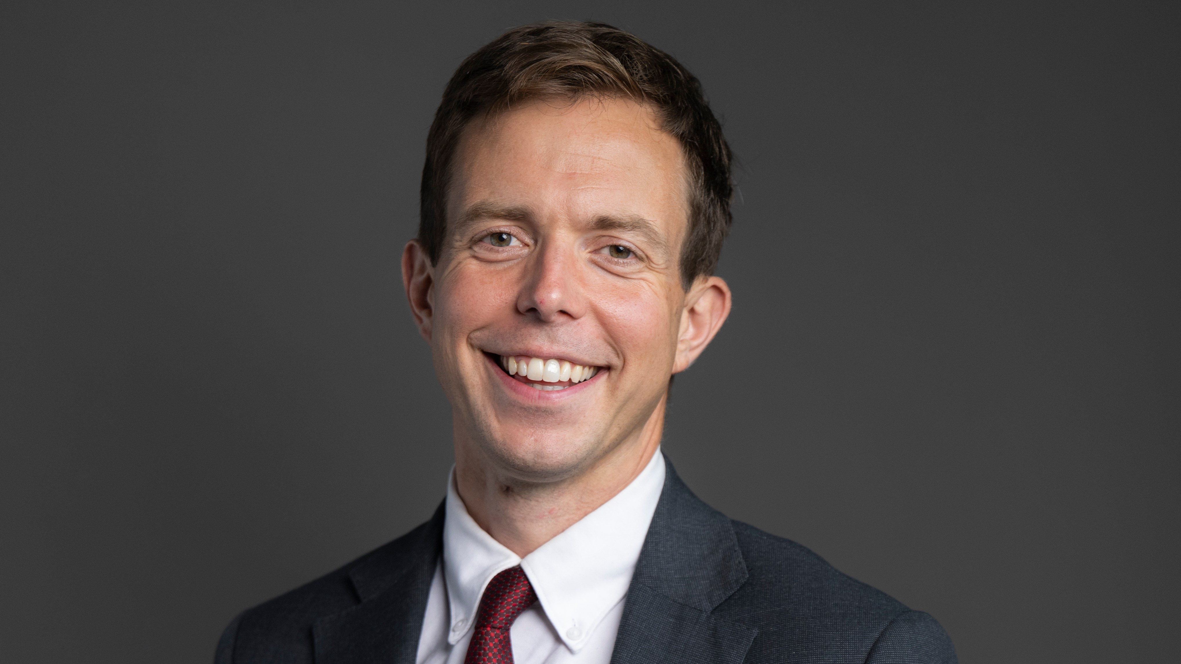 Headshot of Jake Richards MP, a white man in a dark suit and red tie, smiling at the camera.