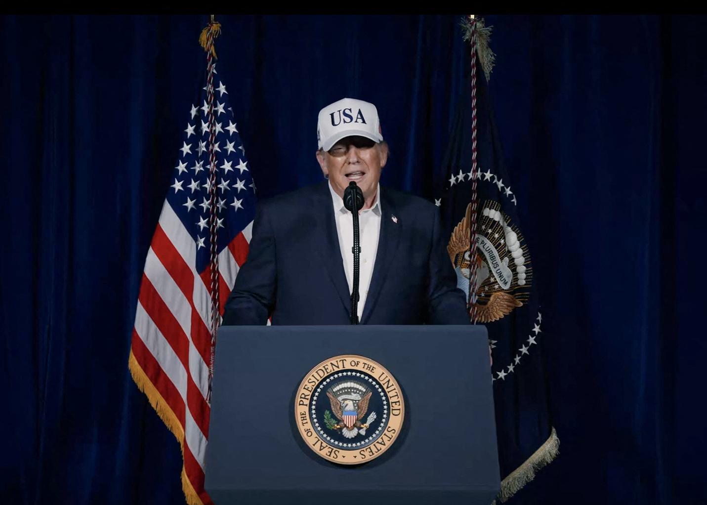 Donald Trump speaking at a podium with a USA hat, flanked by American and presidential flags.