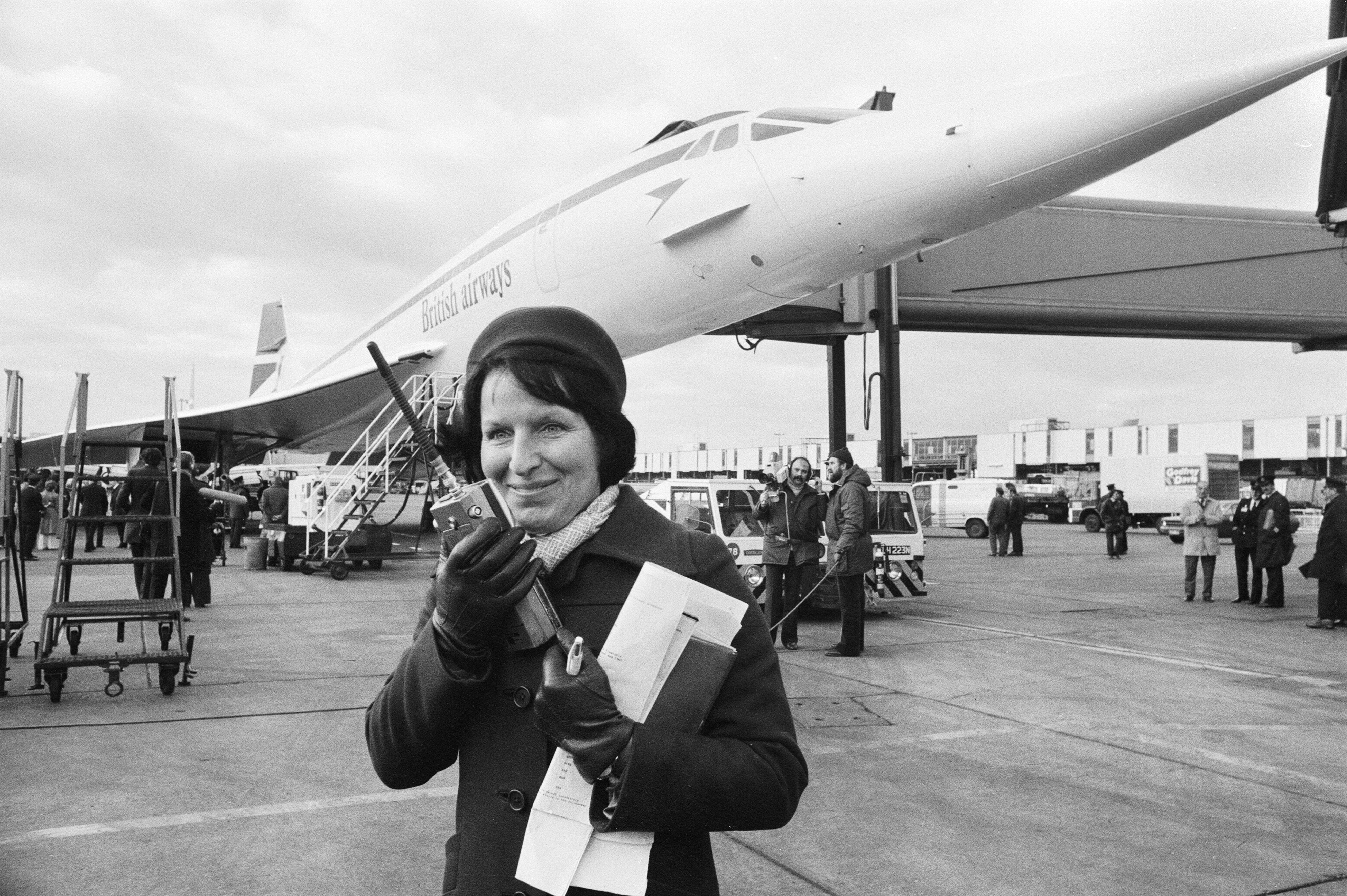 Ramp Liaison Officer Barbara Johnson, holding a walkie-talkie and papers, smiles on the tarmac in front of a British Airways Concorde during its inaugural commercial flight.