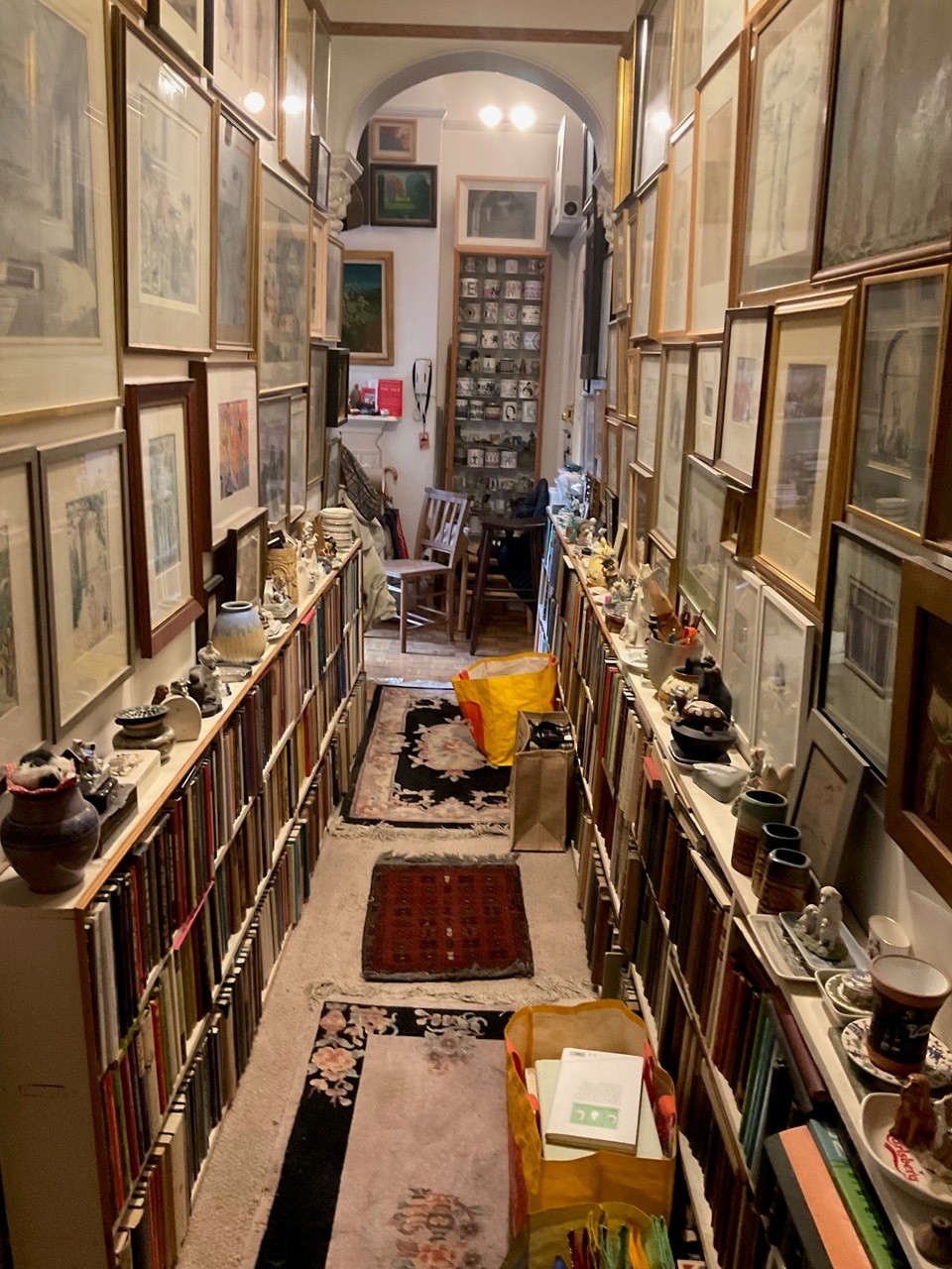 A narrow hall with bookshelves lining both sides and framed art covering the walls, leading to a display cabinet of mugs.